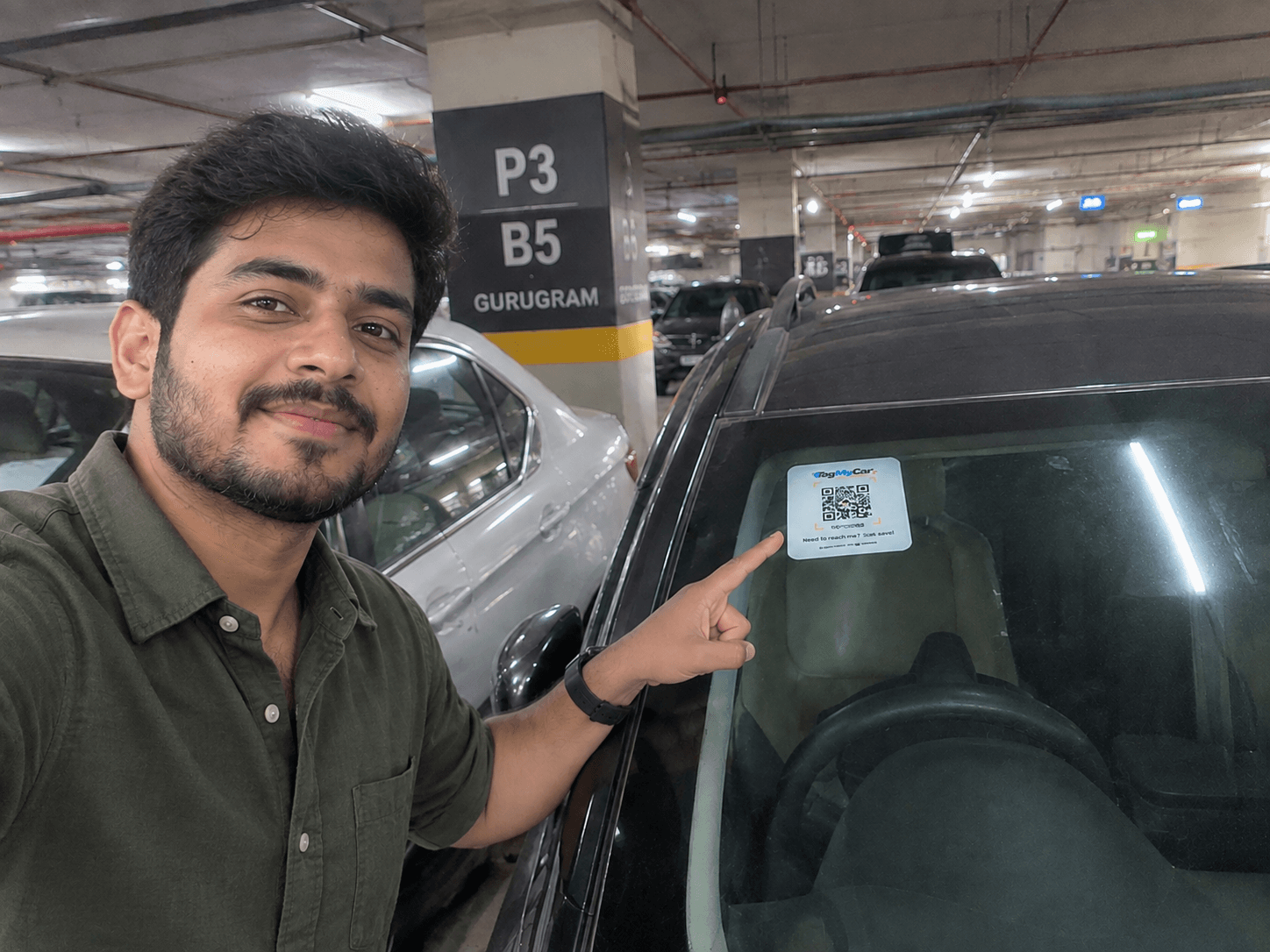Karan, a TagMyCar customer, pointing at the TagMyCar QR tag on his car's rear windshield in a covered parking lot in Gurugram